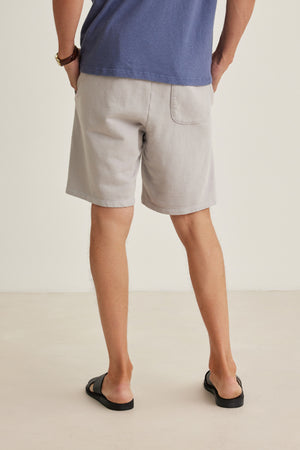 A person standing indoors, seen from behind, wearing a blue t-shirt, Velvet by Graham & Spencer BOSLOW SHORT in light gray with a drawstring waist, black sandals, and a wristwatch.