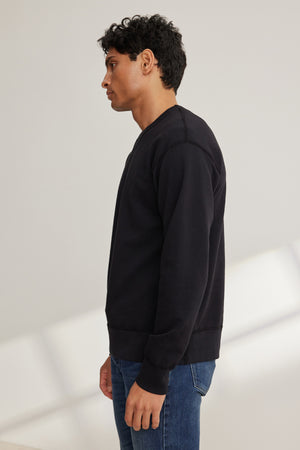 A person in profile wears the RAE SWEATSHIRT by Velvet by Graham & Spencer, a black French terry athletic-inspired top, paired with blue jeans against a simple light background.