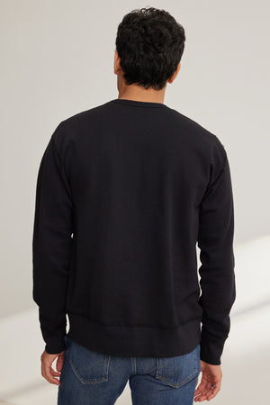 A man faces away from the camera, dressed in Velvet by Graham & Spencer's RAE SWEATSHIRT in black French terry and blue jeans, set against a light background.
