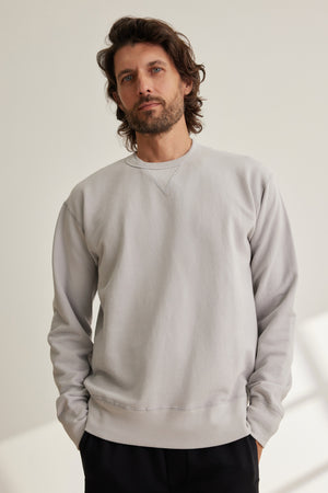 A man with wavy brown hair and a beard stands with hands in pockets, wearing the RAE SWEATSHIRT by Velvet by Graham & Spencer—a light gray premium loopback French terry crewneck—paired with black pants against a plain light background.