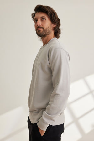 A man with medium-length brown hair and a beard stands indoors, wearing the Velvet by Graham & Spencer RAE SWEATSHIRT in light gray French terry and black pants, looking slightly upward.