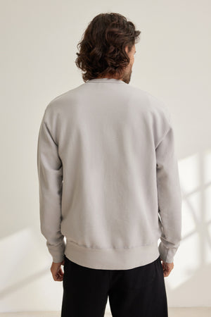 A person with wavy hair wears the Velvet by Graham & Spencer RAE SWEATSHIRT in light gray, featuring a classic crew neckline, and black pants, standing with their back to the camera in a well-lit room.