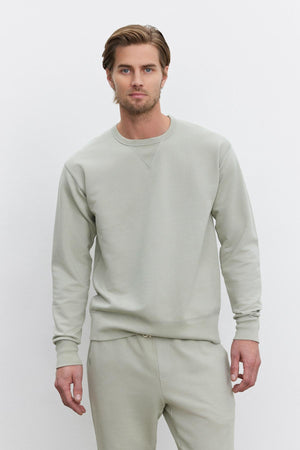 A man with short brown hair and a beard, wearing a light gray Velvet by Graham & Spencer RAE SWEATSHIRT and matching sweatpants, stands against a plain white background.