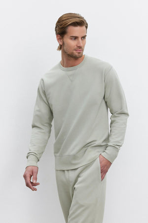 A man with short hair wearing a light grey RAE SWEATSHIRT by Velvet by Graham & Spencer and sweatpants stands against a plain white background.