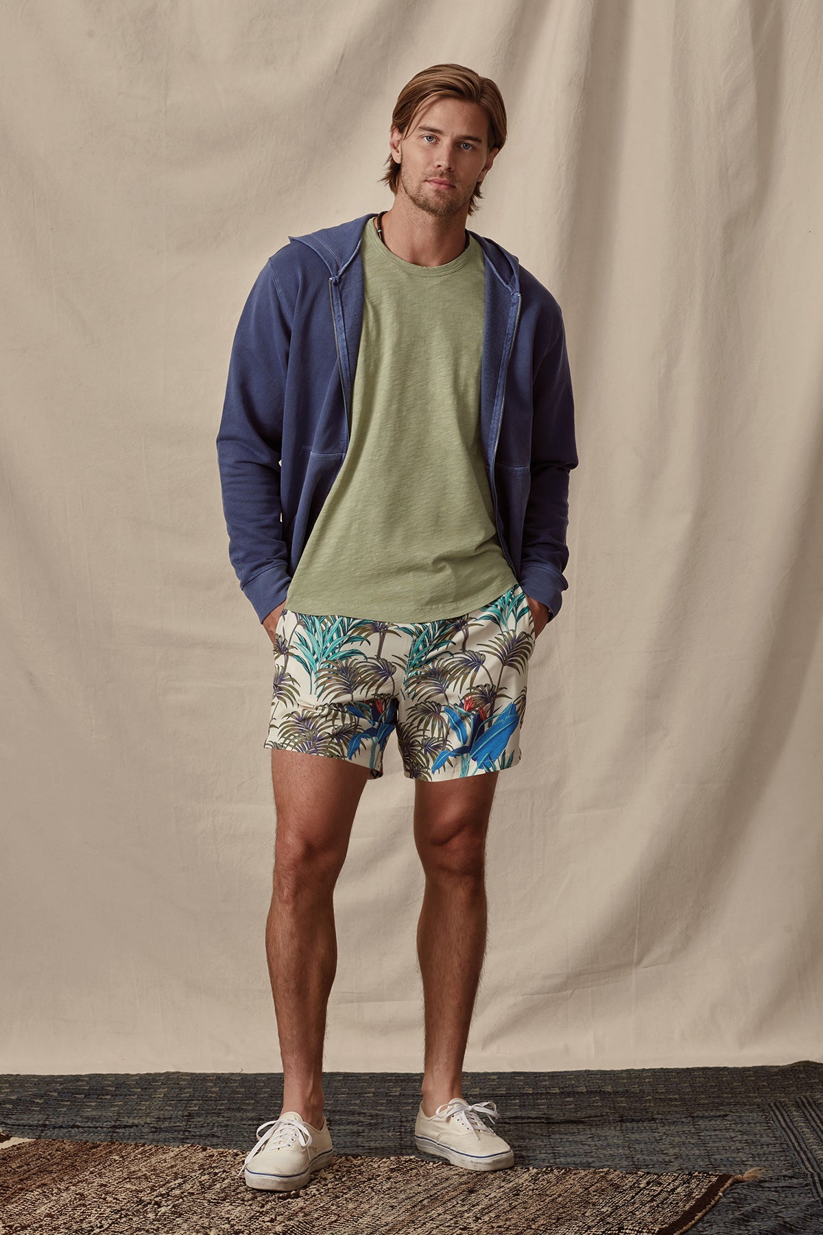   Man standing in a casual pose wearing a navy zip-up hoodie, green crew neck Velvet by Graham & Spencer t-shirt, tropical print shorts, and white sneakers, against a beige backdrop. 