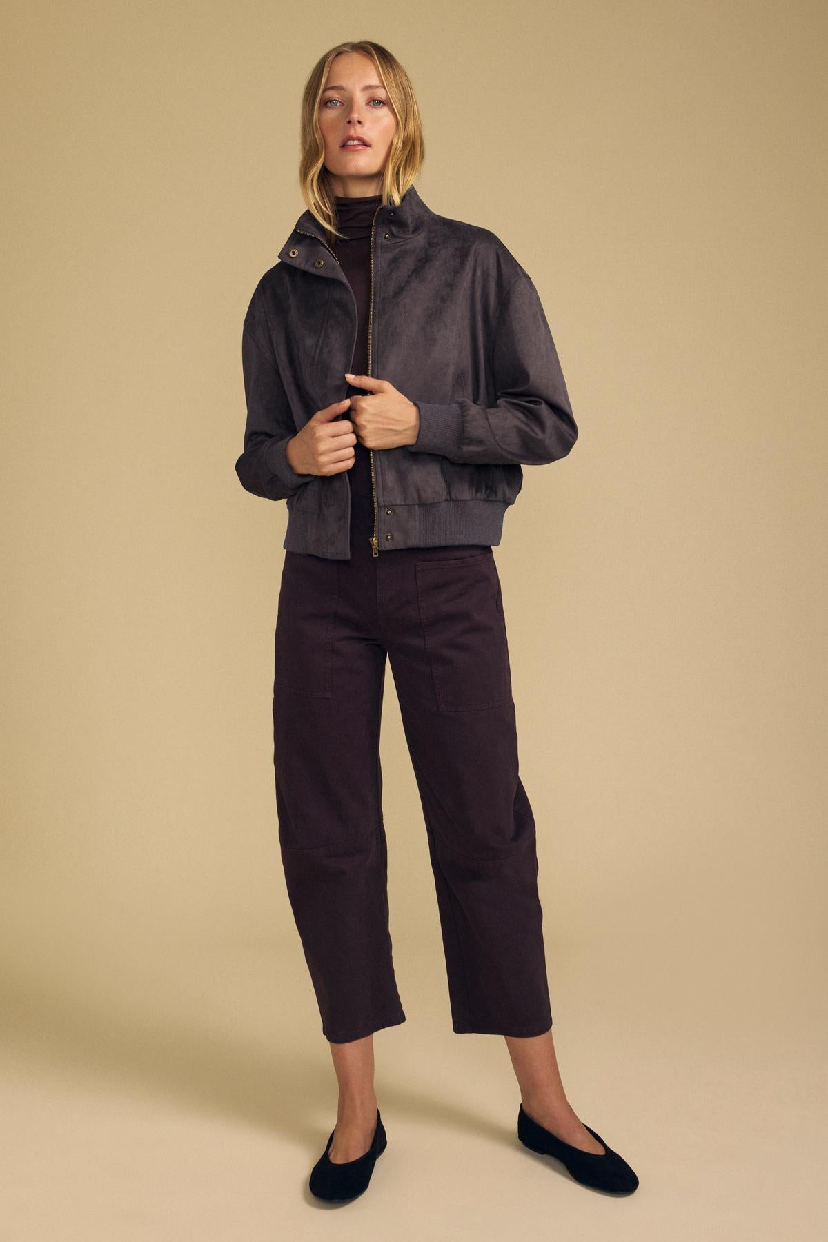 A woman stands against a beige background wearing the QUINCY FAUX SUEDE BOMBER JACKET by Velvet by Graham & Spencer, styled with brown cropped pants, a dark brown turtleneck, and black flats for a modern silhouette.-42157576388801