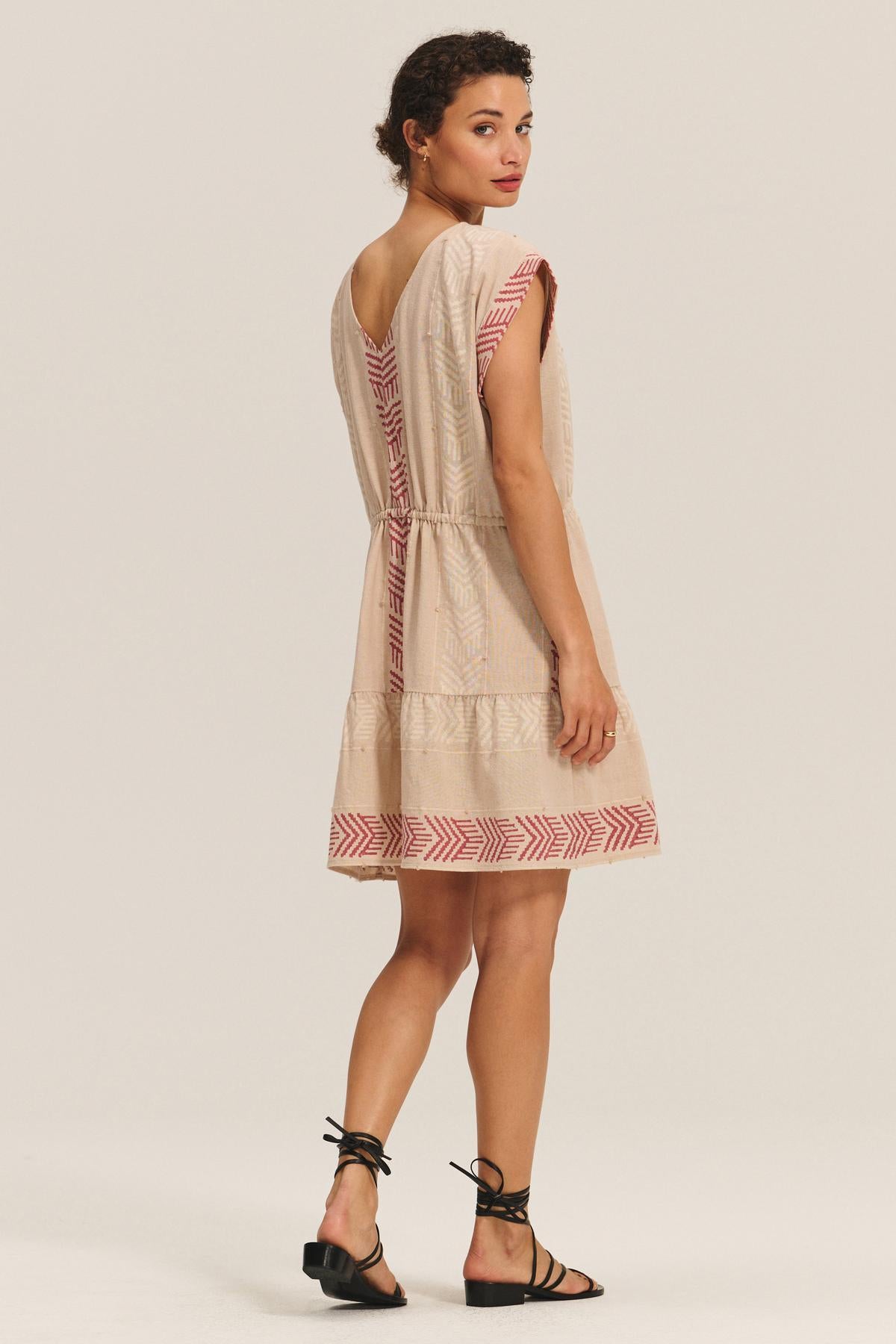 A woman faces away in the CHANTEL DRESS by Velvet by Graham & Spencer, a short-sleeve beige cotton jacquard dress with red geometric patterns, paired with black sandals against a plain light background.-40732758966465