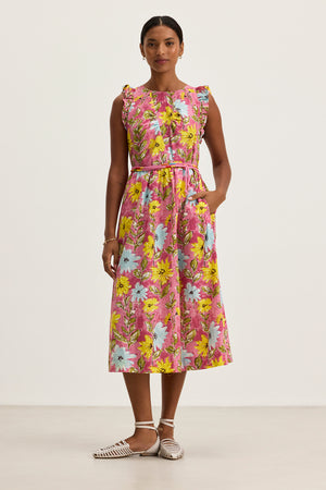 A woman stands in a studio wearing the ELARIA DRESS by Velvet by Graham & Spencer, a sleeveless, ankle-length cotton dress with side seam pockets, a bright pink, yellow, and blue floral print, tied waist, and white woven flats.