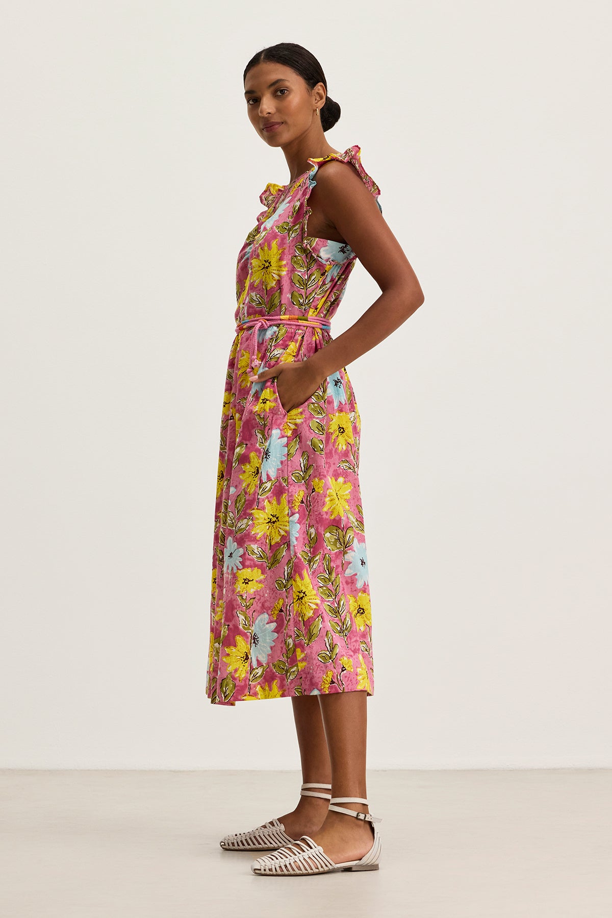 A woman stands sideways against a plain background wearing the ELARIA DRESS by Velvet by Graham & Spencer—a sleeveless, floral cotton midi dress with side seam pockets—paired with flat woven shoes.-39333203837121