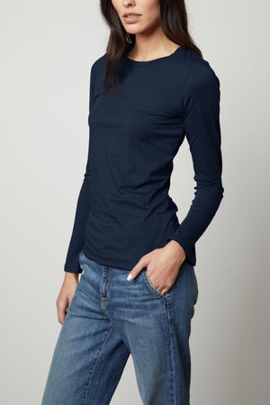 A person with long dark hair wearing a navy blue Velvet by Graham & Spencer ZOFINA TEE and blue jeans stands against a plain white background.