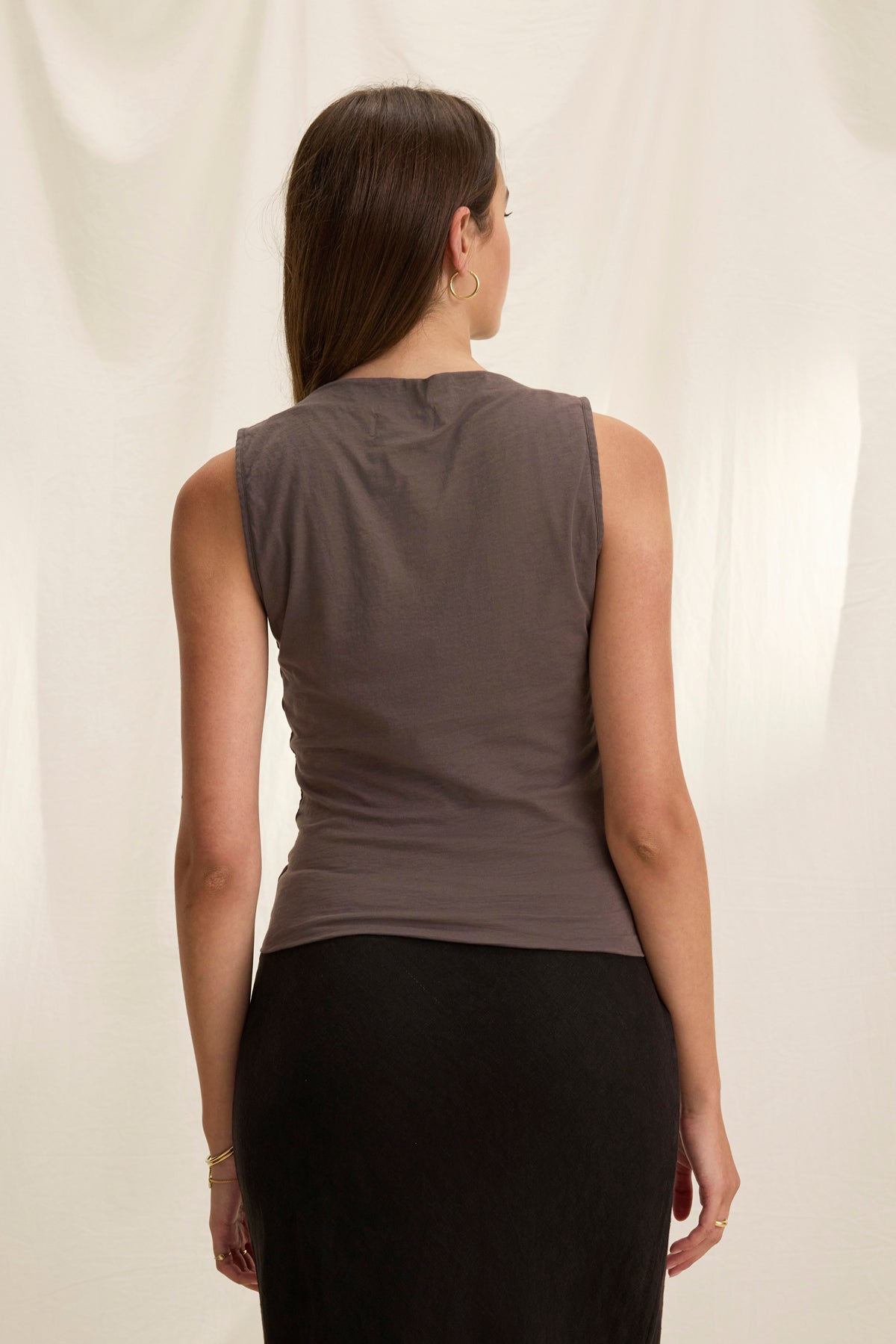 A woman with long hair stands with her back to the camera, wearing the EMILIA TANK TOP from Velvet by Graham & Spencer and a dark skirt. The light background softly highlights the gauzy whisper of her outfit's fabric.-38943210799297