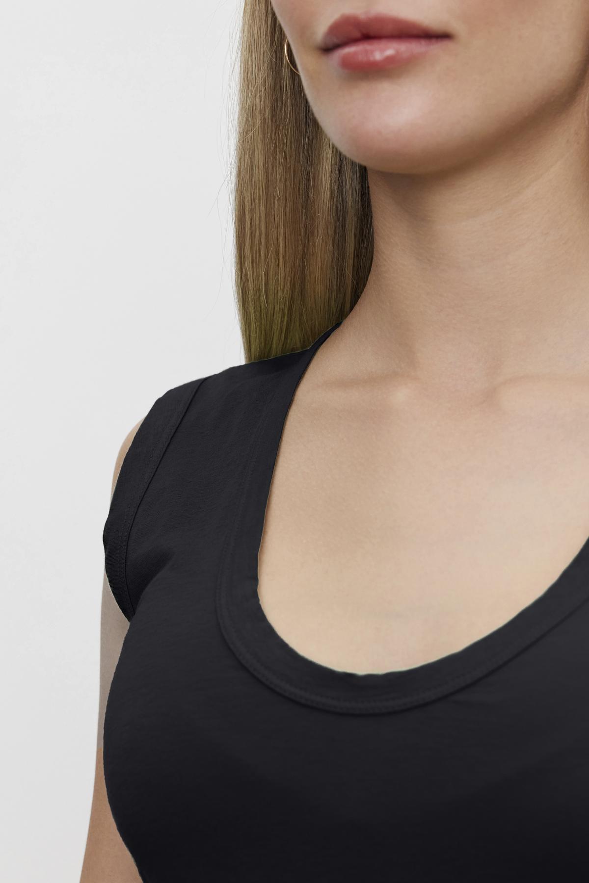 Close-up of a woman wearing a black ESTINA TANK TOP by Velvet by Graham & Spencer, showcasing the neckline and part of her face, with straight, light brunette hair against a plain background.