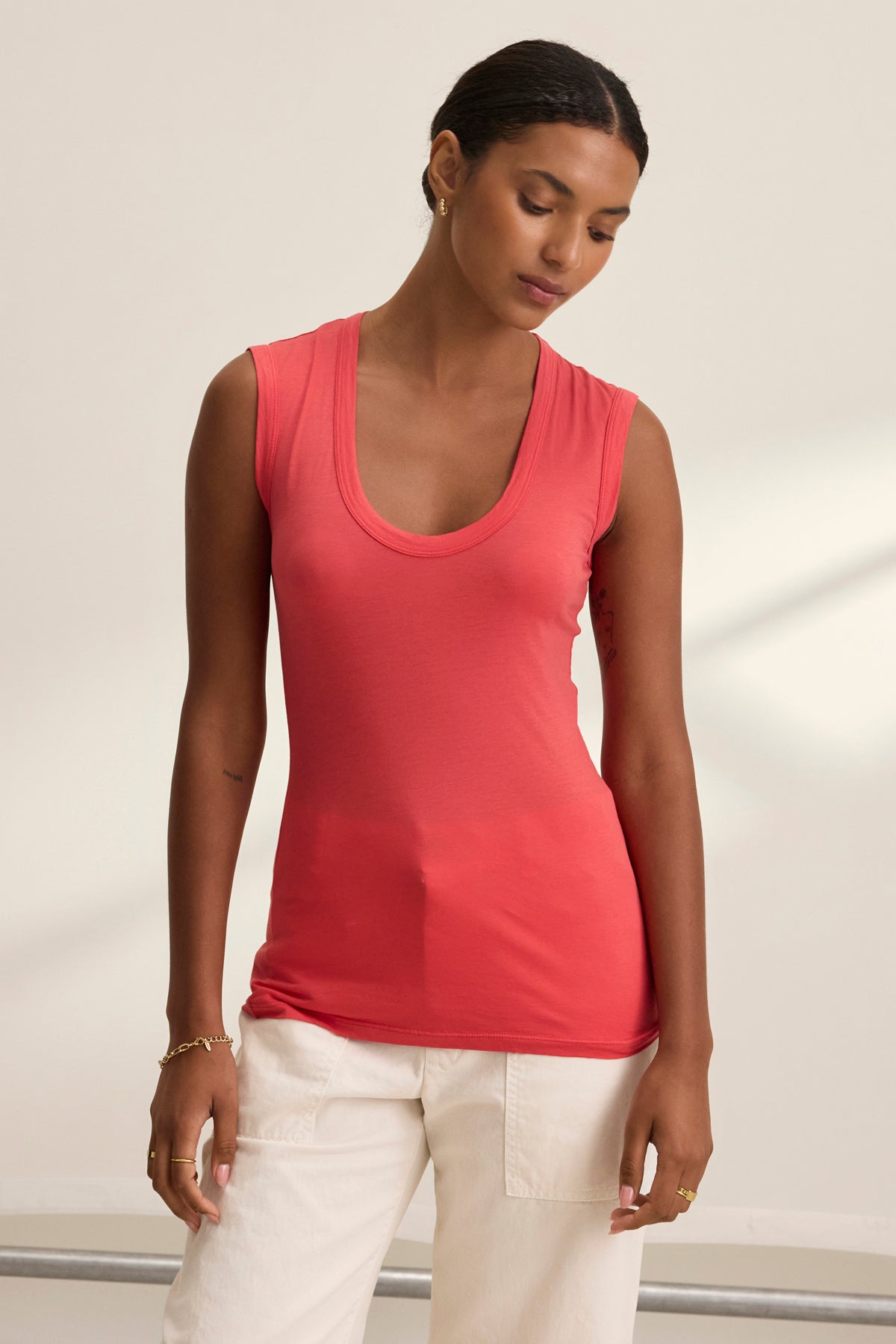 A person wearing the Velvet by Graham & Spencer ESTINA TANK TOP, featuring a scoop-neck design, is paired with white pants and stands against a neutral background, gazing downward.-38928283238593