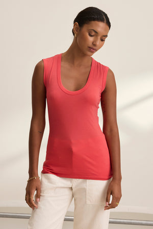 A person wearing the Velvet by Graham & Spencer ESTINA TANK TOP, featuring a scoop-neck design, is paired with white pants and stands against a neutral background, gazing downward.