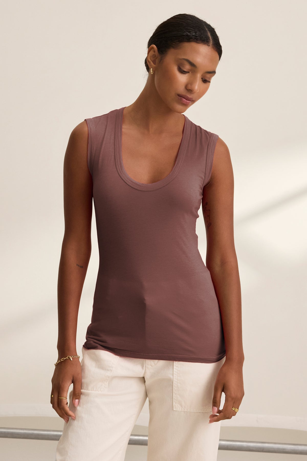A person stands against a plain, light background wearing the ESTINA TANK TOP by Velvet by Graham & Spencer—a scoop-neck, sleeveless mauve top—paired with white pants, looking down with relaxed hands at their sides.-39520708264129