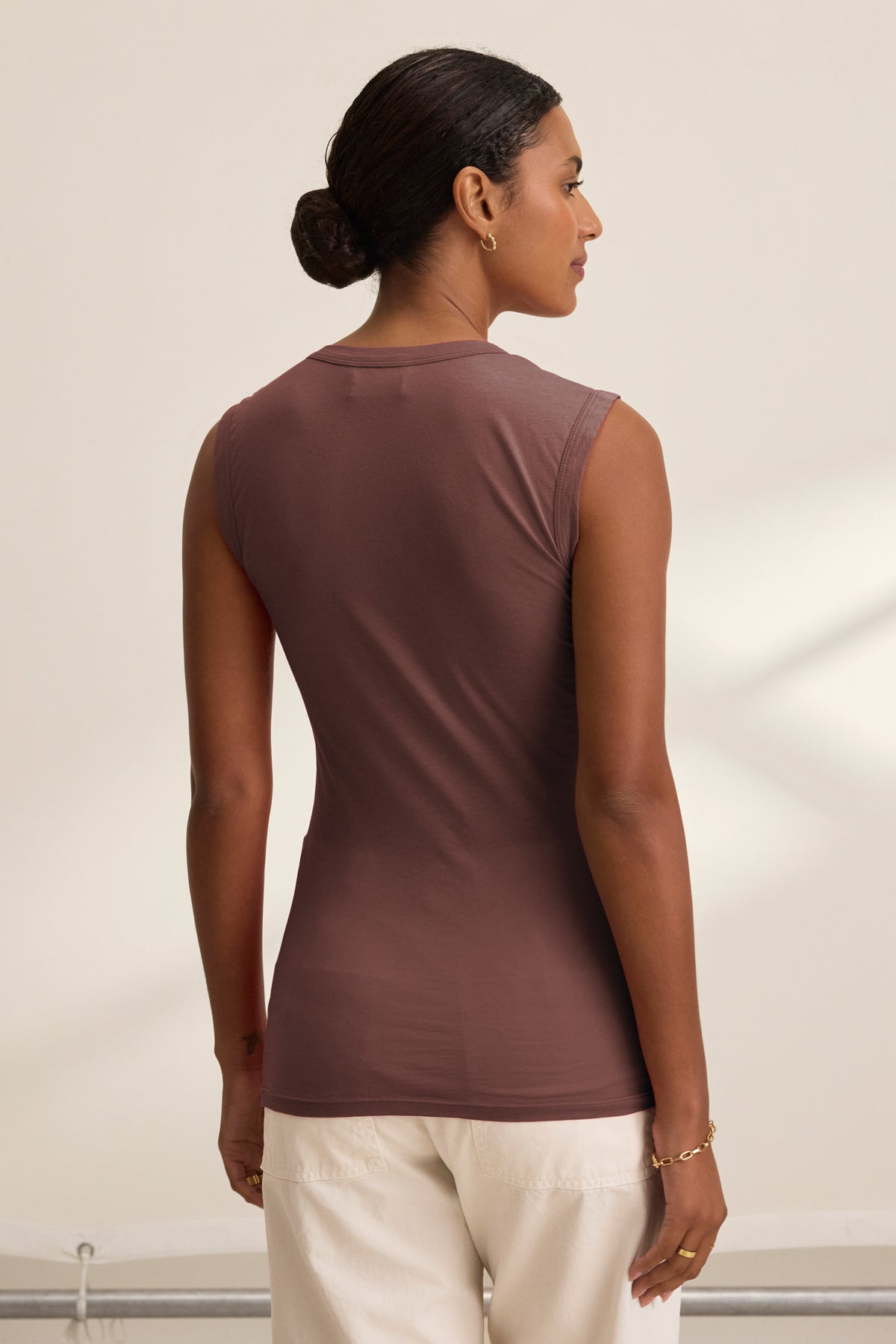 A woman with dark hair in a low bun is seen from the back, wearing the ESTINA TANK TOP by Velvet by Graham & Spencer in brown, paired with white pants against a neutral background.-39520708198593