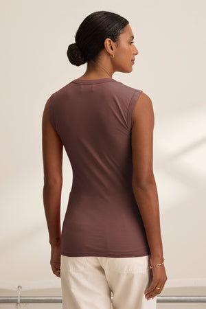 A woman with dark hair in a low bun is seen from the back, wearing the ESTINA TANK TOP by Velvet by Graham & Spencer in brown, paired with white pants against a neutral background.