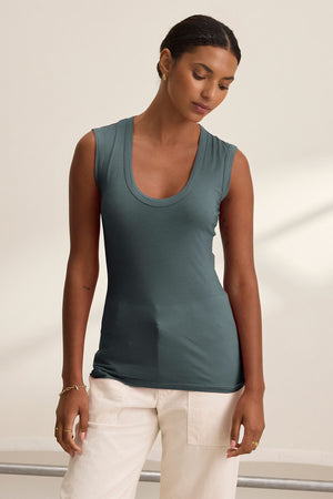 Wearing the ESTINA TANK TOP by Velvet by Graham & Spencer in teal, paired with white pants, a person stands indoors against a plain light-colored wall.