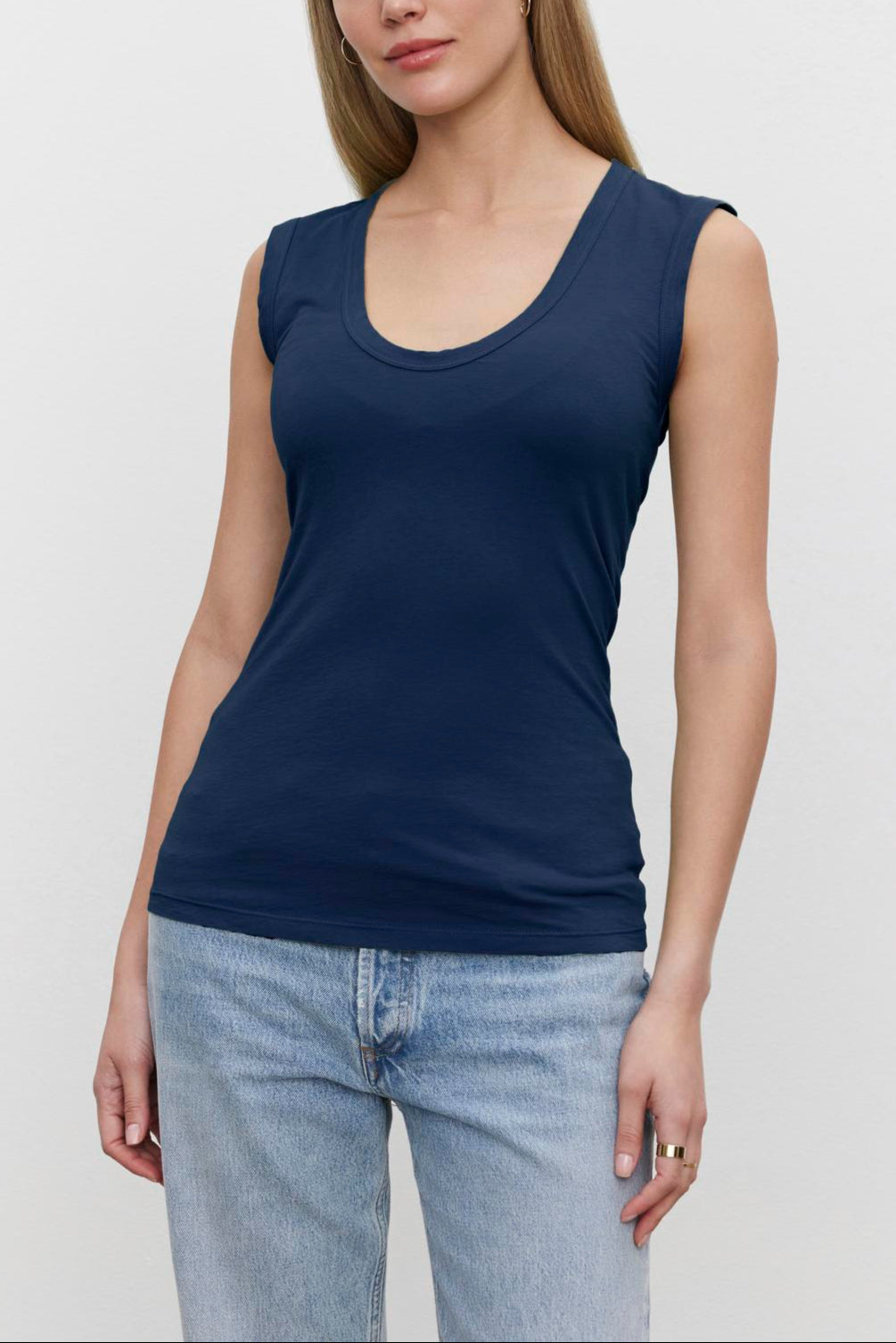 A woman with long hair is wearing a navy blue low-scoop-neck sleeveless Velvet by Graham & Spencer ESTINA TANK TOP and light blue jeans, standing against a plain white background.