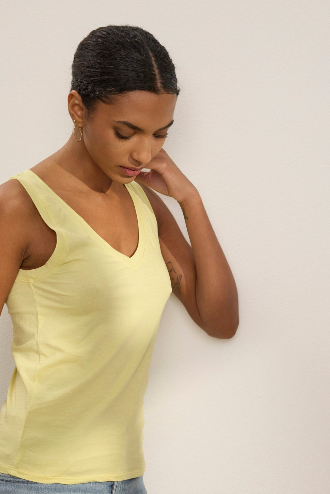Wearing the EVELYN TANK TOP by Velvet by Graham & Spencer, a person stands against a light-colored wall, looking down with one hand touching their neck.