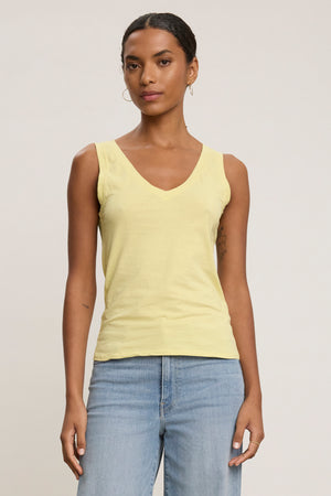 A woman models the EVELYN TANK TOP by Velvet by Graham & Spencer—a light yellow, sleeveless V-neck piece—paired with light blue jeans against a plain background, highlighting its versatility as a wardrobe essential.
