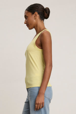 Woman in profile wears the EVELYN TANK TOP by Velvet by Graham & Spencer in light yellow, paired with blue jeans and gold hoops, hair in a neat bun against a plain backdrop—a true wardrobe essential.