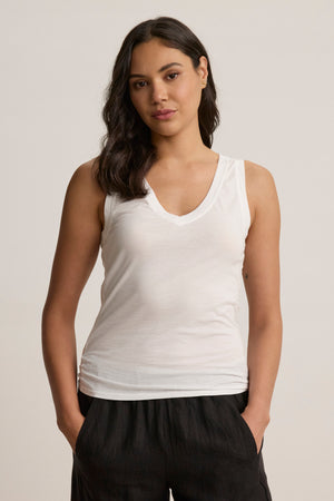 A woman with long dark hair wears the EVELYN TANK TOP by Velvet by Graham & Spencer— a versatile white sleeveless V-neck tank—paired with black pants, standing hands-in-pockets against a plain background.