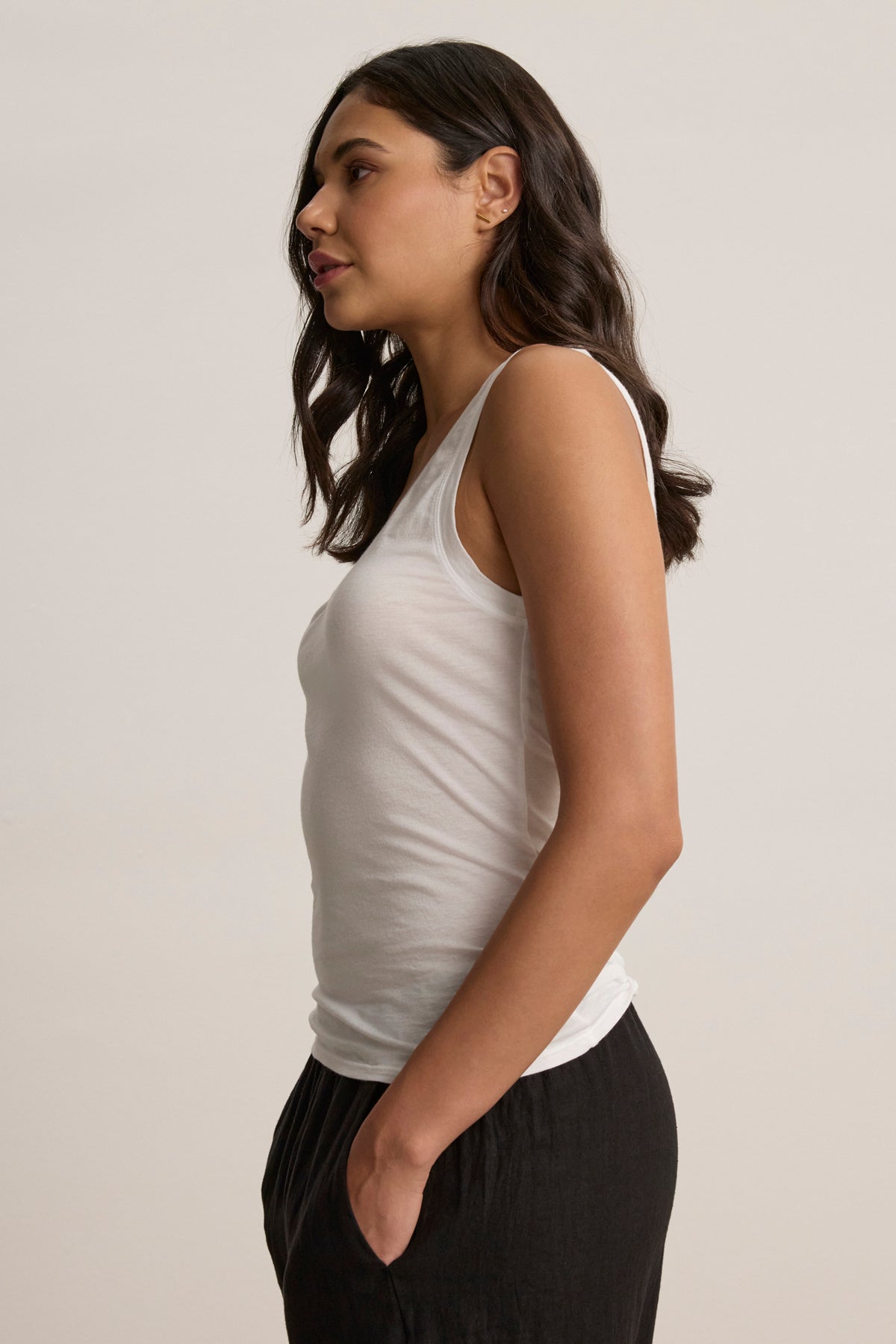 A woman with long dark hair stands in profile, wearing the EVELYN TANK TOP by Velvet by Graham & Spencer and black pants, her hand in her pocket against a plain background.