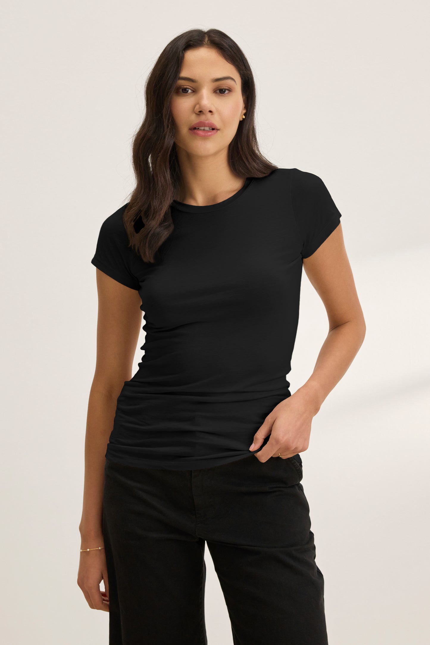 A woman with long dark hair wears the JEMMA TEE by Velvet by Graham & Spencer—a fitted, ultra-soft crew neck tee—paired with black pants, standing against a plain light background.-40183209296065