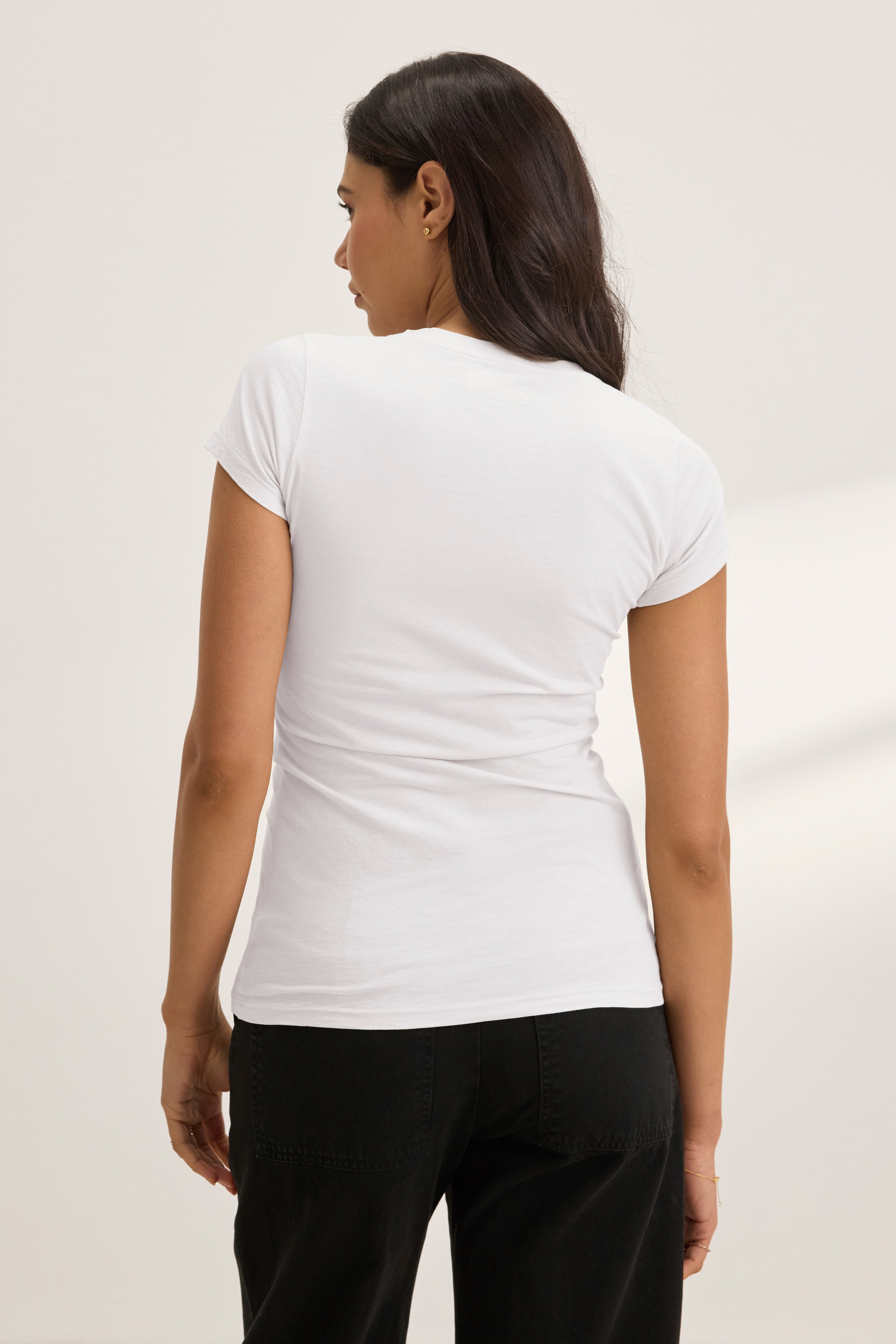 A woman with long dark hair stands with her back to the camera against a plain background, wearing the JEMMA TEE by Velvet by Graham & Spencer in white paired with black pants.