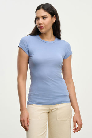 A woman with long dark hair wears the JEMMA TEE by Velvet by Graham & Spencer, an ultra-soft light blue fitted top, paired with beige pants against a plain light background.