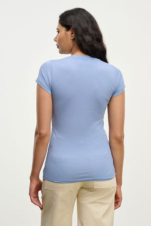 A woman with long dark hair wears the JEMMA TEE by Velvet by Graham & Spencer in light blue, paired with beige pants, standing with her back to the camera against a plain background.