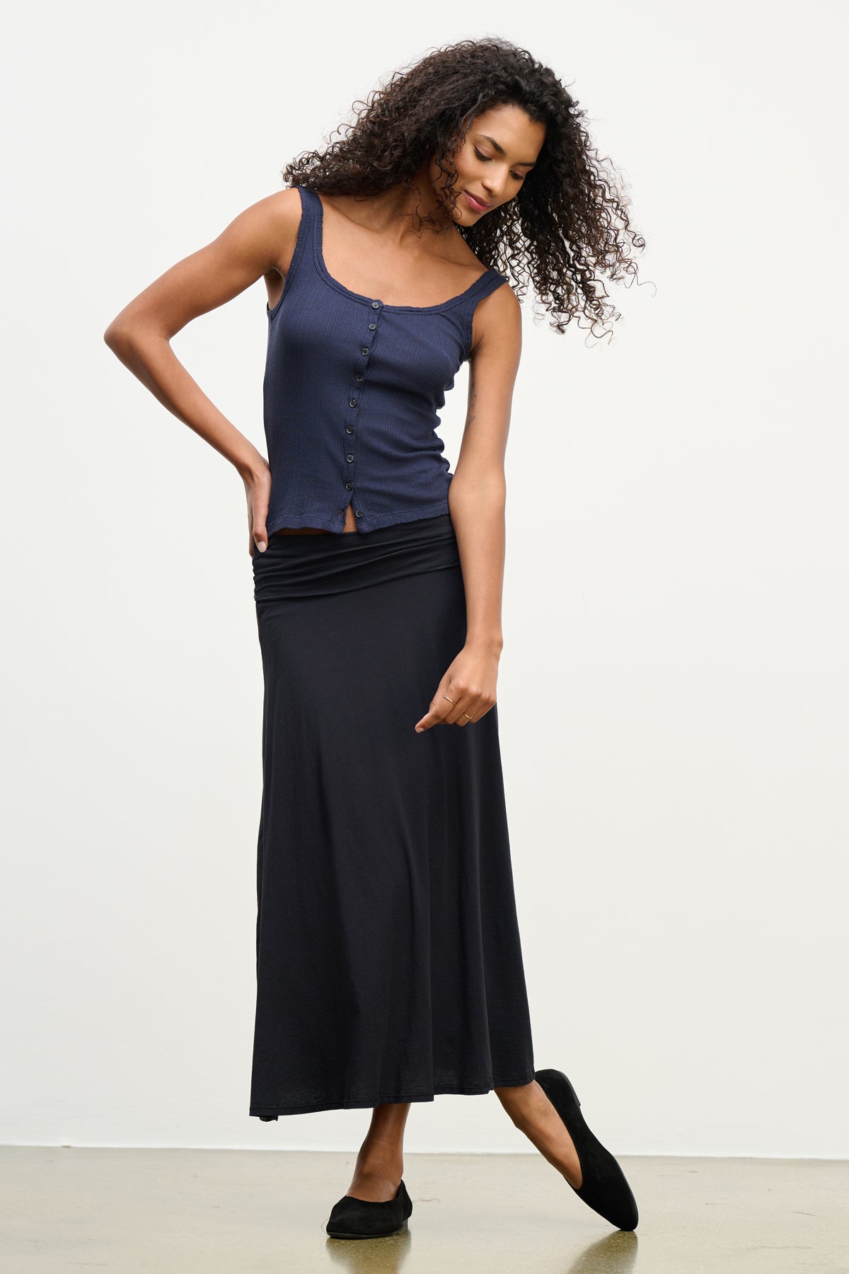 A woman stands on a light floor wearing the LUELLA SKIRT by Velvet by Graham & Spencer—a long black A-line skirt—paired with a navy sleeveless buttoned top and black flats, her head tilted and one hand touching her skirt.
