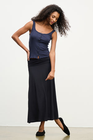A woman stands on a light floor wearing the LUELLA SKIRT by Velvet by Graham & Spencer—a long black A-line skirt—paired with a navy sleeveless buttoned top and black flats, her head tilted and one hand touching her skirt.