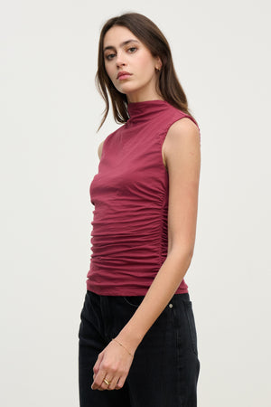 A woman wears the MADELINE TANK TOP by Velvet by Graham & Spencer—a sleeveless maroon mock neck style—paired with black jeans, standing against a plain light background and gazing slightly toward the camera.