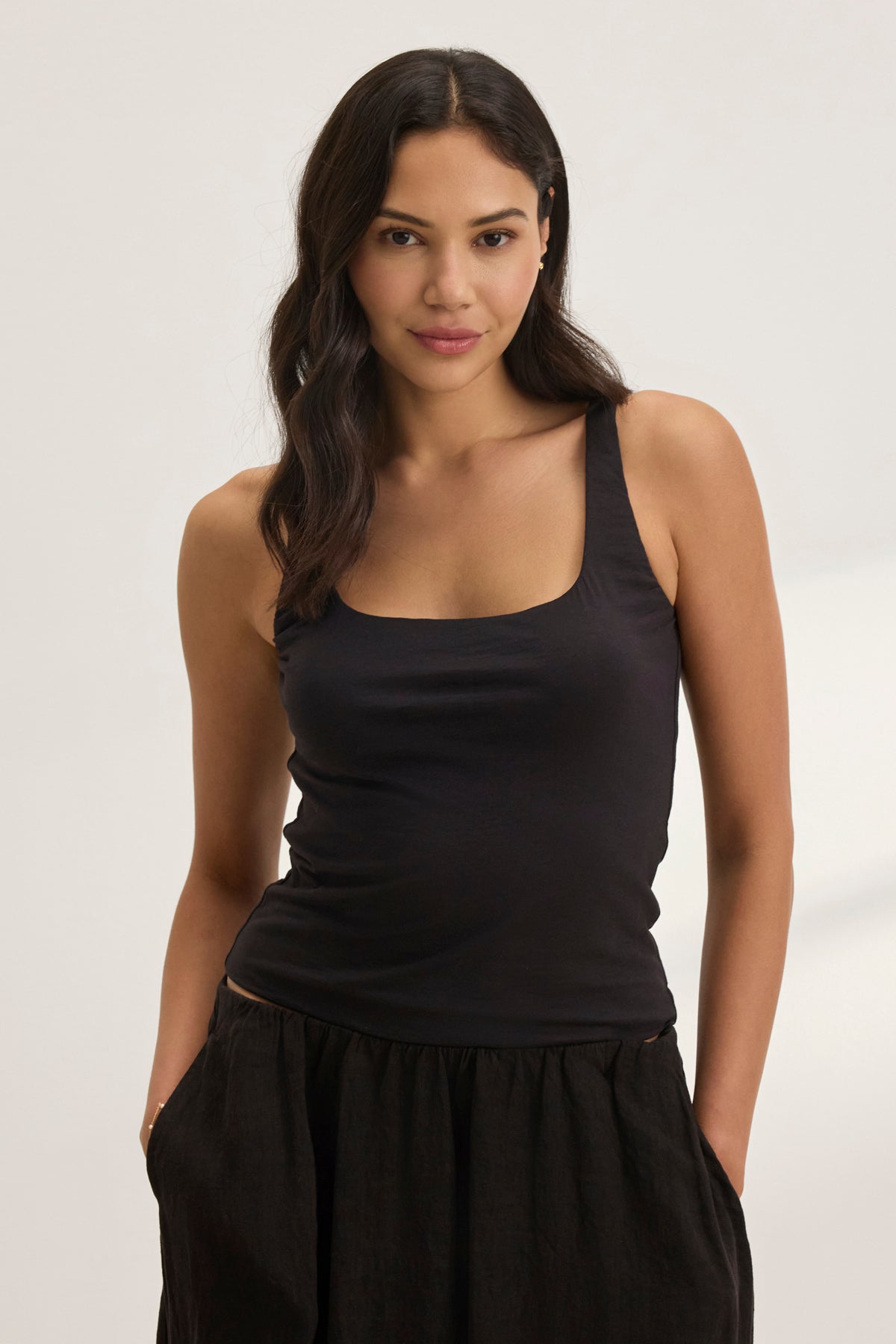 A person with long dark hair stands against a plain background, hands in pockets, wearing the MELANI TANK TOP by Velvet by Graham & Spencer—a black tank featuring a scoop neckline—and dark pants.-38957715783873