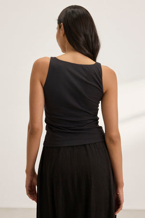 A long-haired individual stands with their back to the camera in a well-lit room, wearing a black MELANI TANK TOP by Velvet by Graham & Spencer and a skirt.