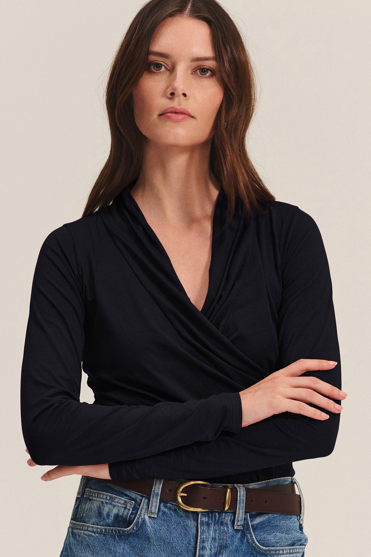 A woman with long brown hair, wearing Velvet by Graham & Spencer’s MERI TEE, blue jeans, and a brown belt, stands with arms crossed against a plain background.