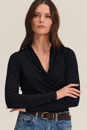 A woman with long brown hair, wearing Velvet by Graham & Spencer’s MERI TEE, blue jeans, and a brown belt, stands with arms crossed against a plain background.