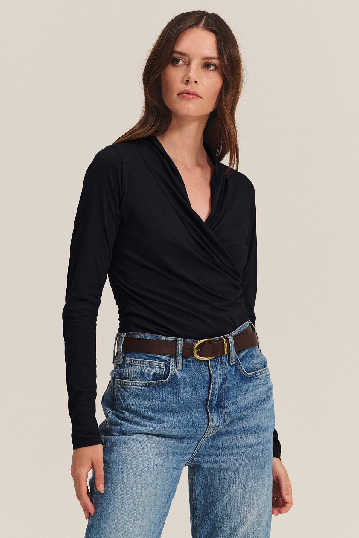 A woman stands against a plain light background wearing the Velvet by Graham & Spencer MERI TEE, a black long-sleeve wrap top in gauzy whisper fabric, tucked into blue jeans with a brown belt.