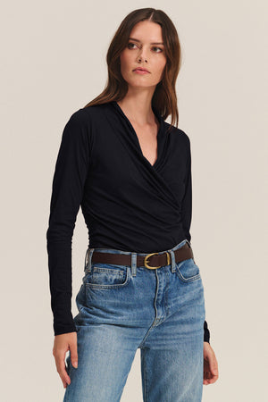 A woman stands against a plain light background wearing the Velvet by Graham & Spencer MERI TEE, a black long-sleeve wrap top in gauzy whisper fabric, tucked into blue jeans with a brown belt.
