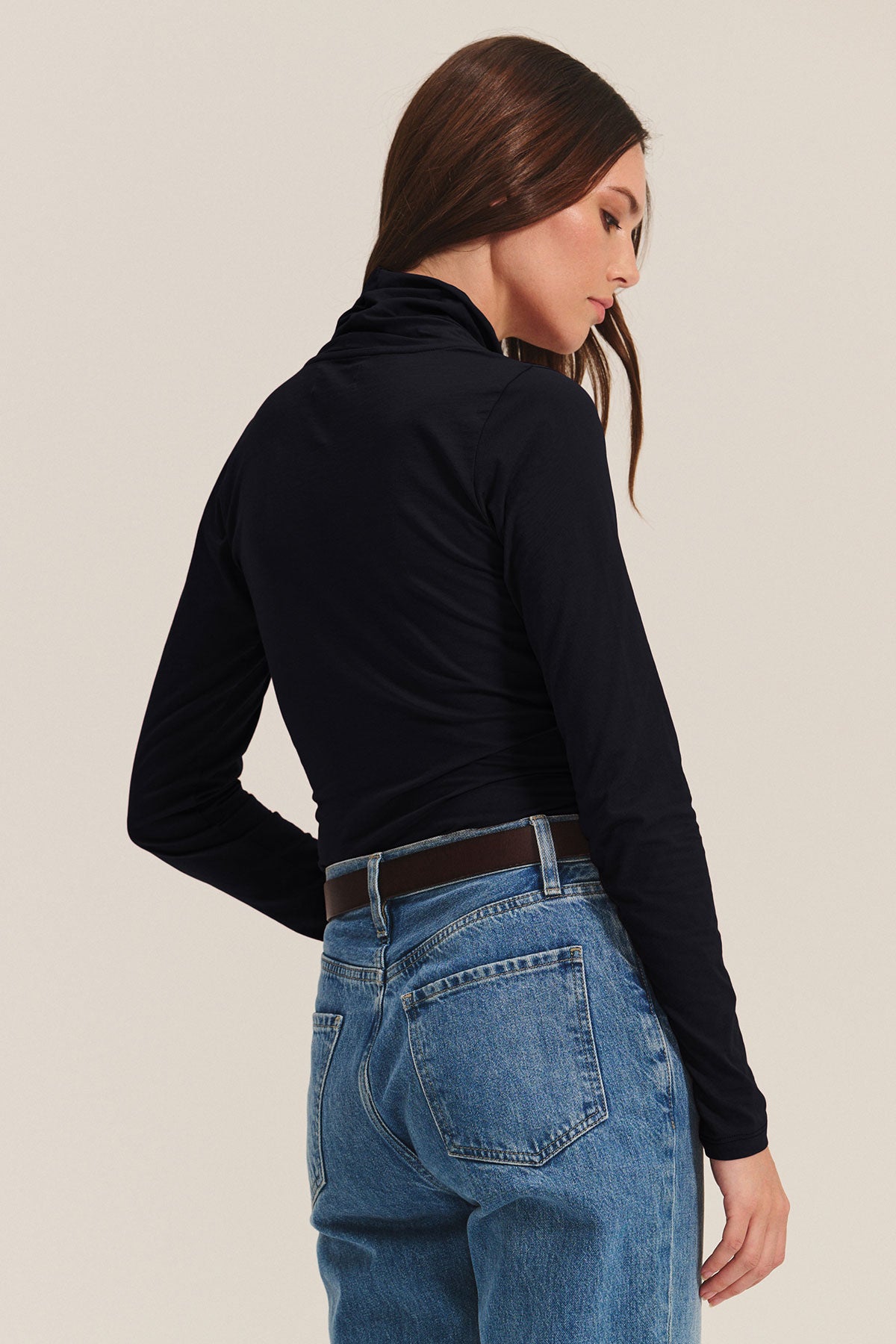 A woman stands with her back to the camera, wearing the MERI TEE black long-sleeve turtleneck by Velvet by Graham & Spencer paired with blue jeans.