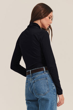 A woman stands with her back to the camera, wearing the MERI TEE black long-sleeve turtleneck by Velvet by Graham & Spencer paired with blue jeans.