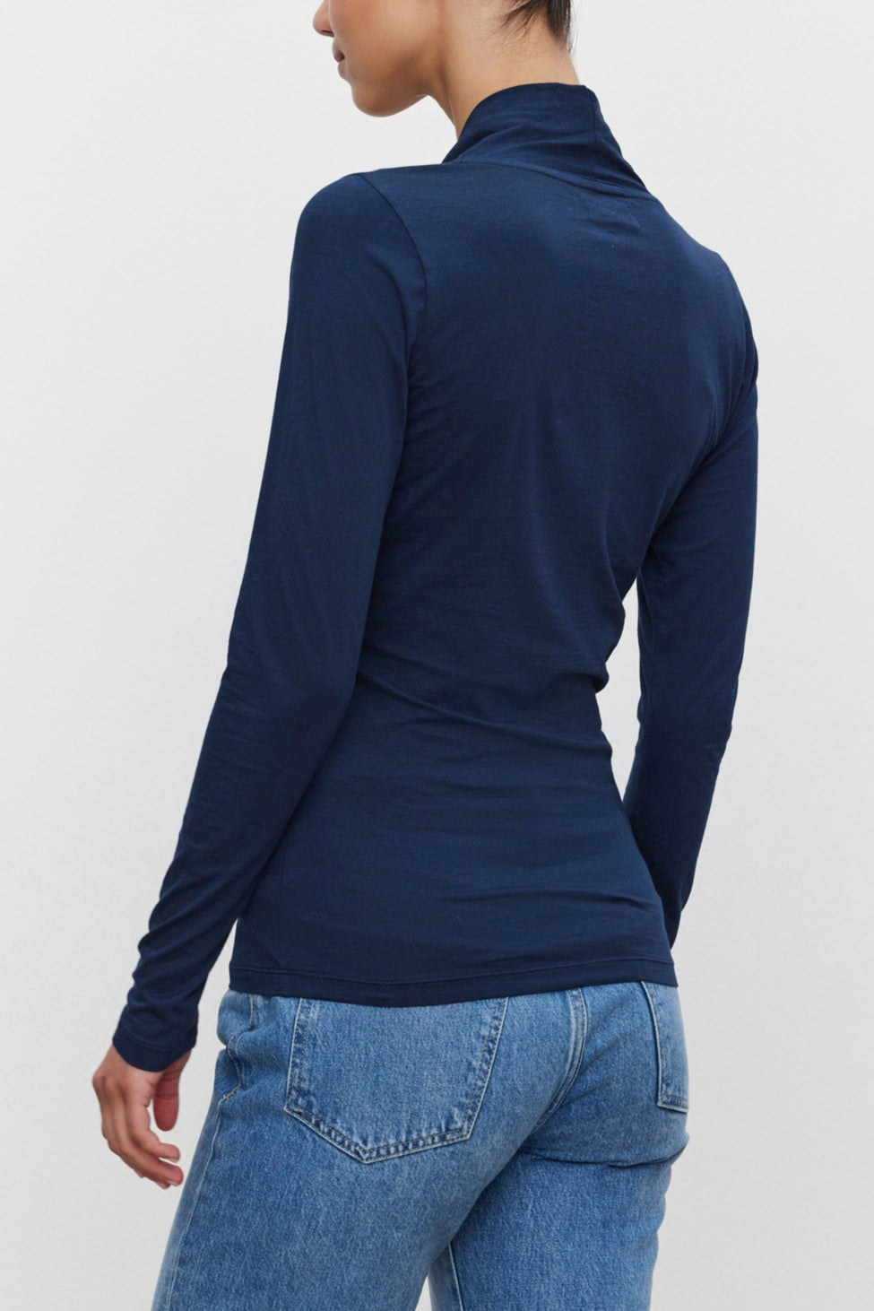 A person with dark hair is seen from behind, sporting a fitted silhouette in the MERI TEE long-sleeved, navy blue top by Velvet by Graham & Spencer and blue jeans. The hair is styled in a neat bun.-37856500711617