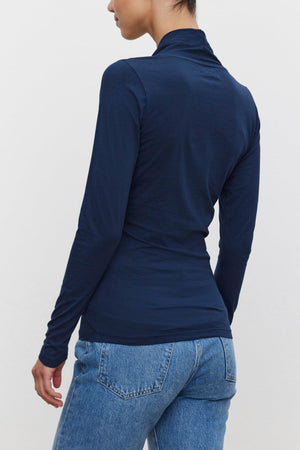 A person with dark hair is seen from behind, sporting a fitted silhouette in the MERI TEE long-sleeved, navy blue top by Velvet by Graham & Spencer and blue jeans. The hair is styled in a neat bun.