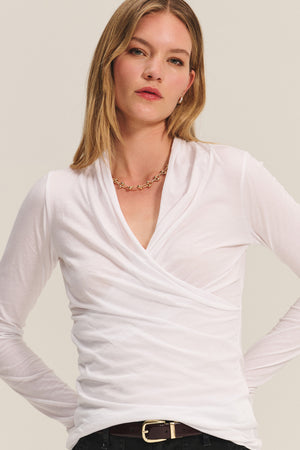 A woman with straight blonde hair wears the MERI TEE by Velvet by Graham & Spencer, styled with a gold chain necklace and black belted pants, standing against a neutral background.