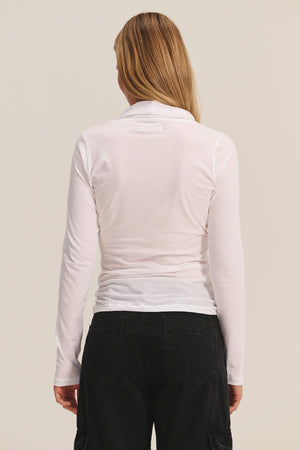 A person with long blonde hair is shown from the back, wearing the MERI TEE by Velvet by Graham & Spencer—a light pink fitted V-neck t-shirt—and black pants against a plain background.
