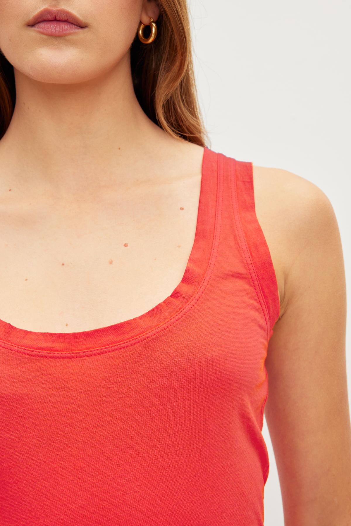 Close-up of a person wearing a MOSSY TANK TOP in red by Velvet by Graham & Spencer, featuring minimal jewelry with gold hoop earrings. The background is a plain white wall, highlighting the versatile wardrobe choice.-36010151346369