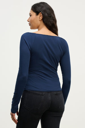 A woman with long dark hair, wearing the Velvet by Graham & Spencer RAK TEE and black jeans, stands with her back to the camera against a plain background, showcasing a feminine silhouette.