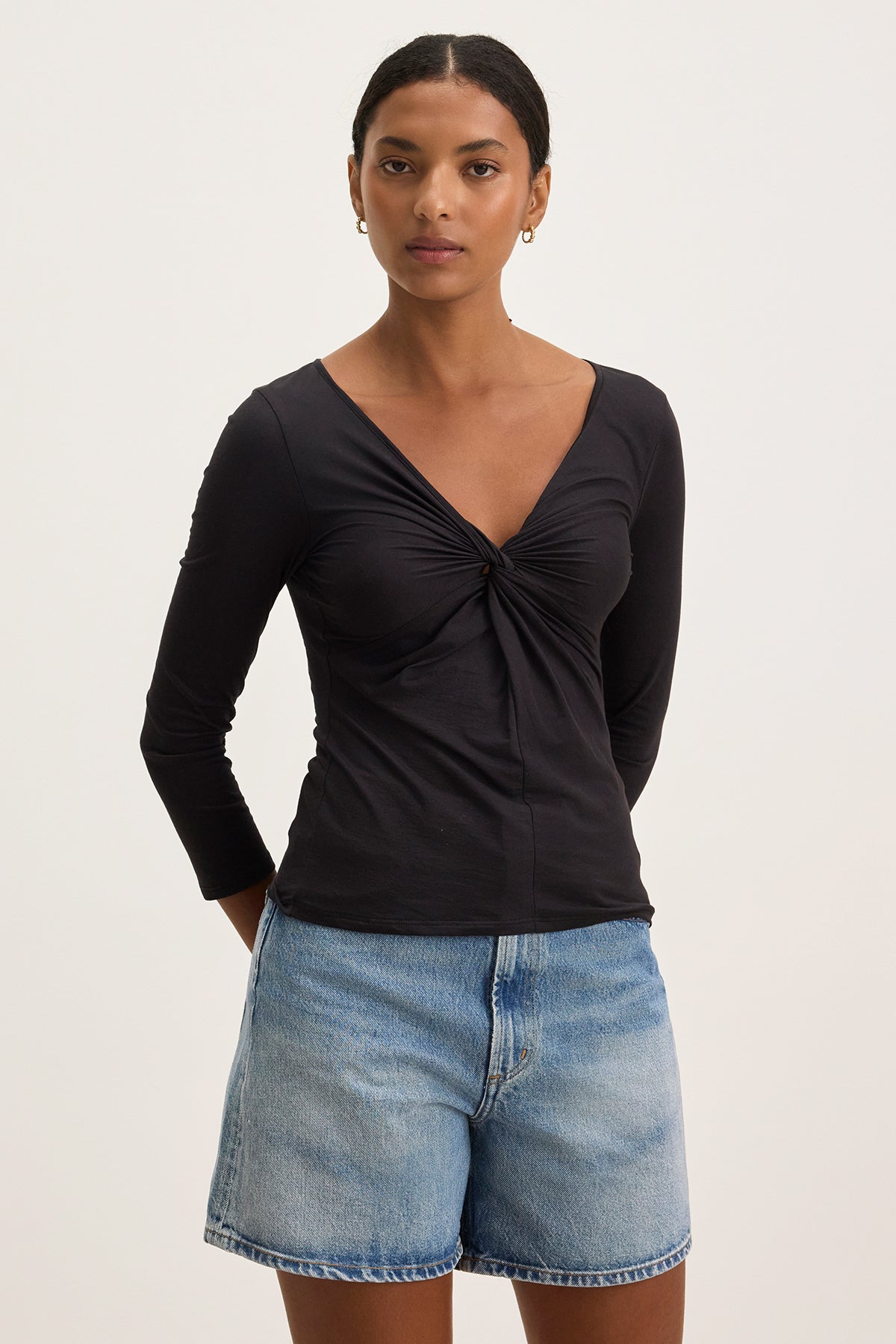 A person confidently stands against a plain background, wearing the SETT TEE by Velvet by Graham & Spencer, a black v-neckline long-sleeve top paired with denim shorts.